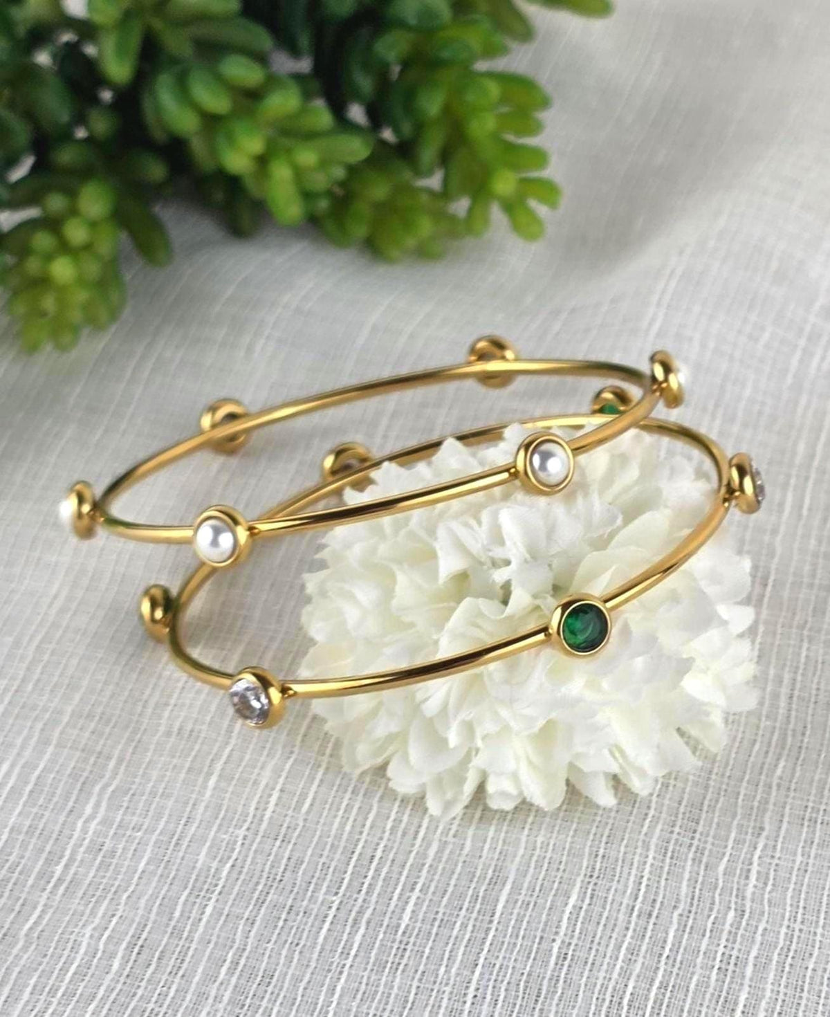 May include: Two gold-colored bangles with decorative accents. One bangle features pearls, a clear gem, and a green gem. The other bangle has pearls and clear gems. The bangles are resting on a white flower.