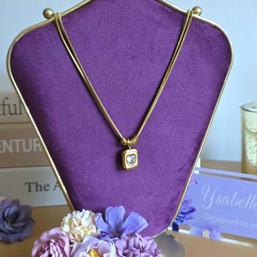 A gold-toned necklace featuring a thin snake chain and a rectangular pendant with a central heart-shaped clear stone. The pendant is suspended from a series of small gold rings, creating a layered look on a purple display stand.