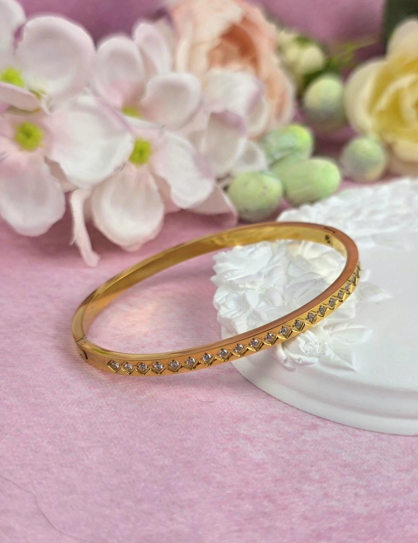 A gold-colored bangle bracelet with a row of small, clear gemstones. The bracelet is set against a soft pink background with white floral accents. The bracelet is a classic piece of jewelry.