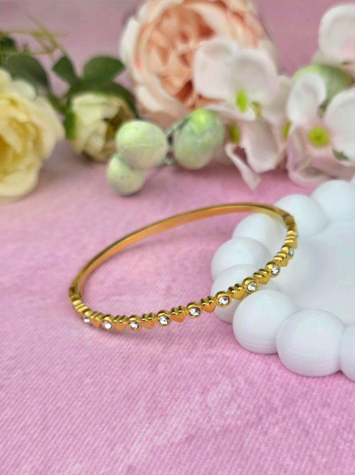 A gold-toned bangle bracelet featuring a repeating pattern of small heart shapes alternating with clear, round, sparkling stones. The metal band has a polished finish and a circular shape, showcasing a delicate and consistent design throughout.
