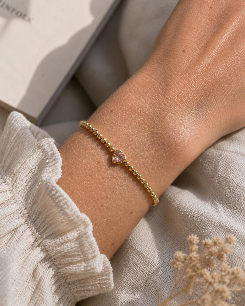 Image includes a  Gold beaded bracelet with a pink heart-shaped charm. The bracelet is worn on a wrist, with a cream-colored sleeve in the background. The bracelet is made of small, round gold beads and the heart charm is a light pink color.