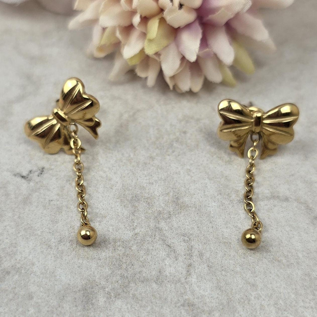 A pair of gold-toned stud earrings featuring a polished bow design. Each earring has a delicate chain dangling from the center of the bow, which terminates in a small, smooth gold-toned sphere at the end of the chain.