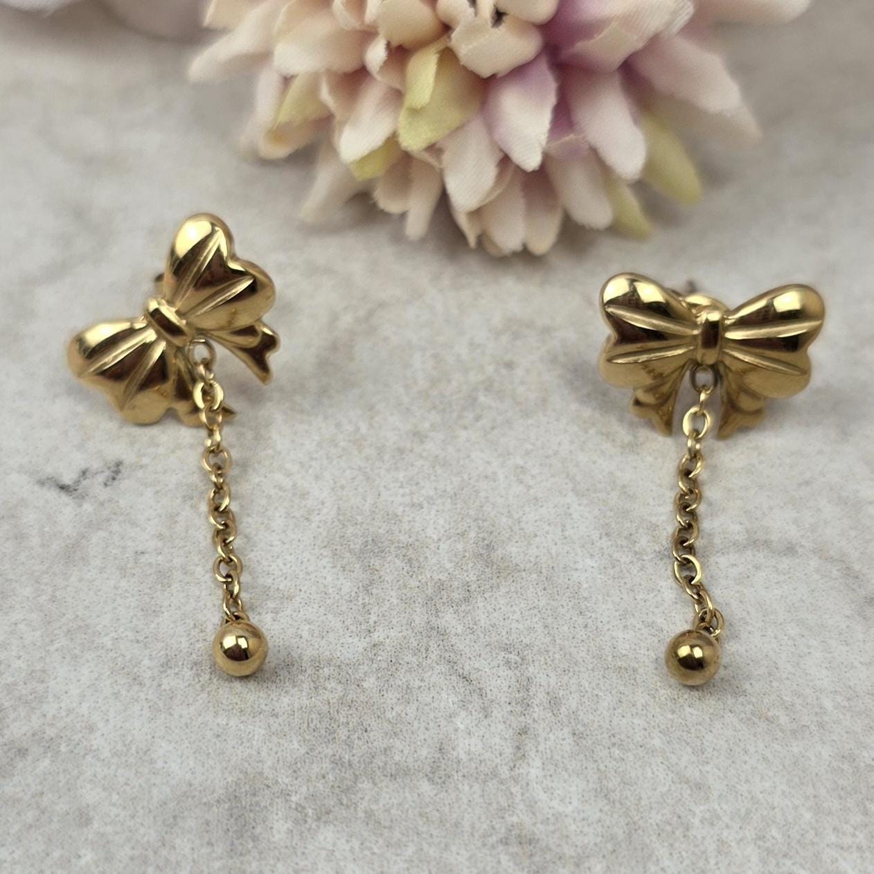 A pair of gold-toned stud earrings featuring a polished bow design. Each earring has a delicate chain dangling from the center of the bow, which terminates in a small, smooth gold-toned sphere at the end of the chain.