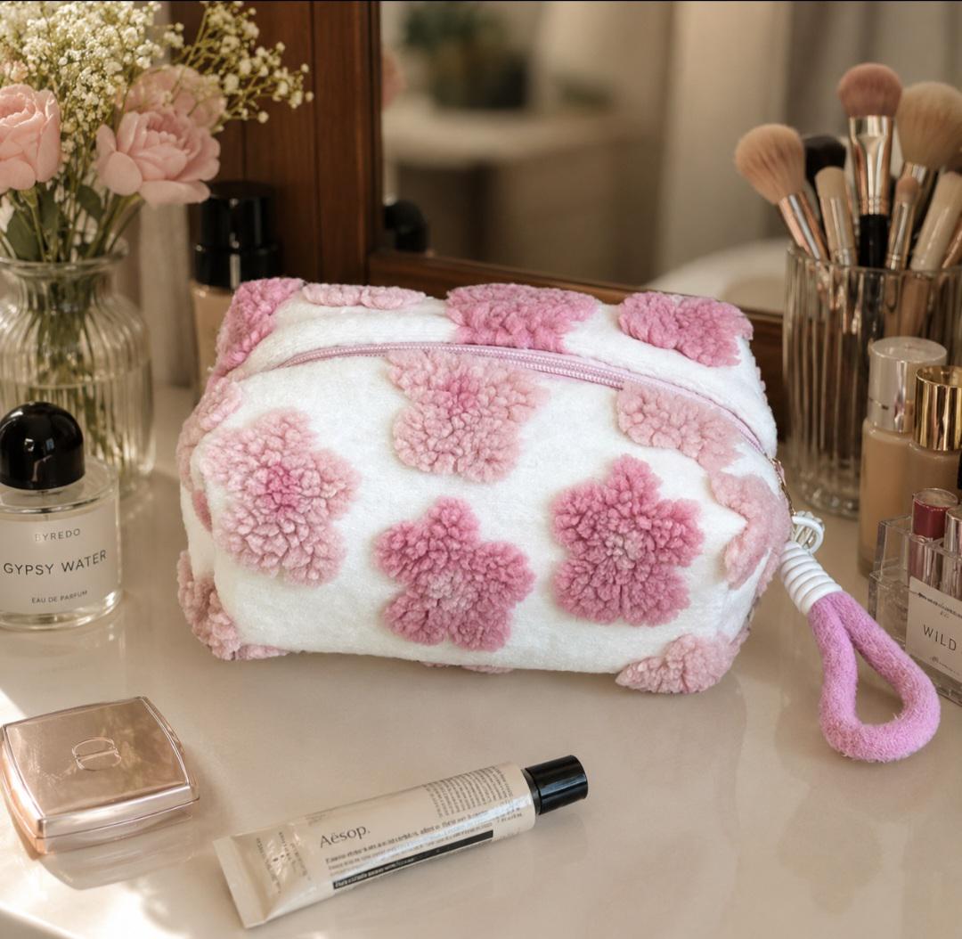 White cosmetic bag with pink flower-shaped accents. The bag has a zipper closure and a pink loop handle. The bag is sitting on a white surface with other beauty products and a vase of flowers.