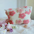 Images include A white cosmetic bag with pink flower-shaped tufted accents. The bag has a rectangular shape and a zipper closure. The bag is placed on a fluffy white surface.