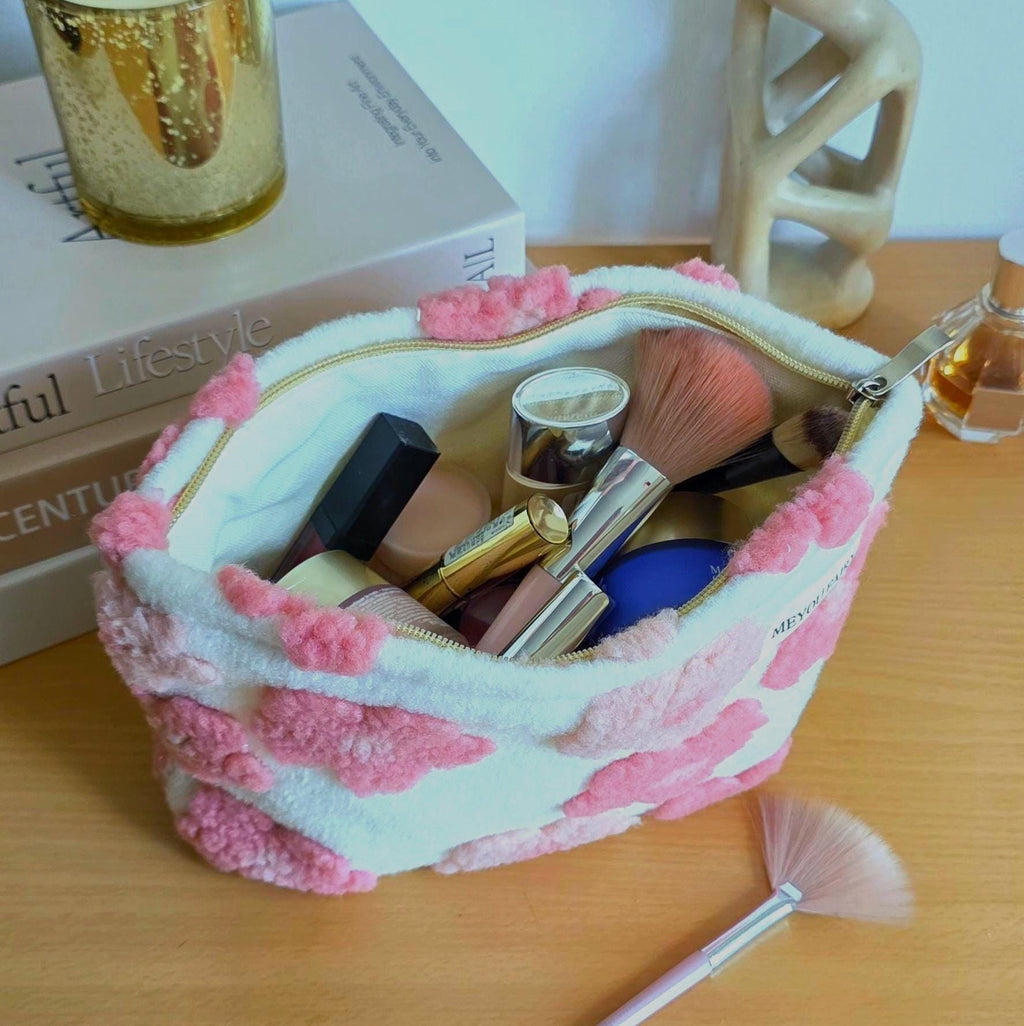 A white and pink patterned cosmetic bag filled with makeup brushes, lipsticks, and other beauty products. The bag has a gold zipper and is sitting on a wooden surface. A fan brush is in the foreground.