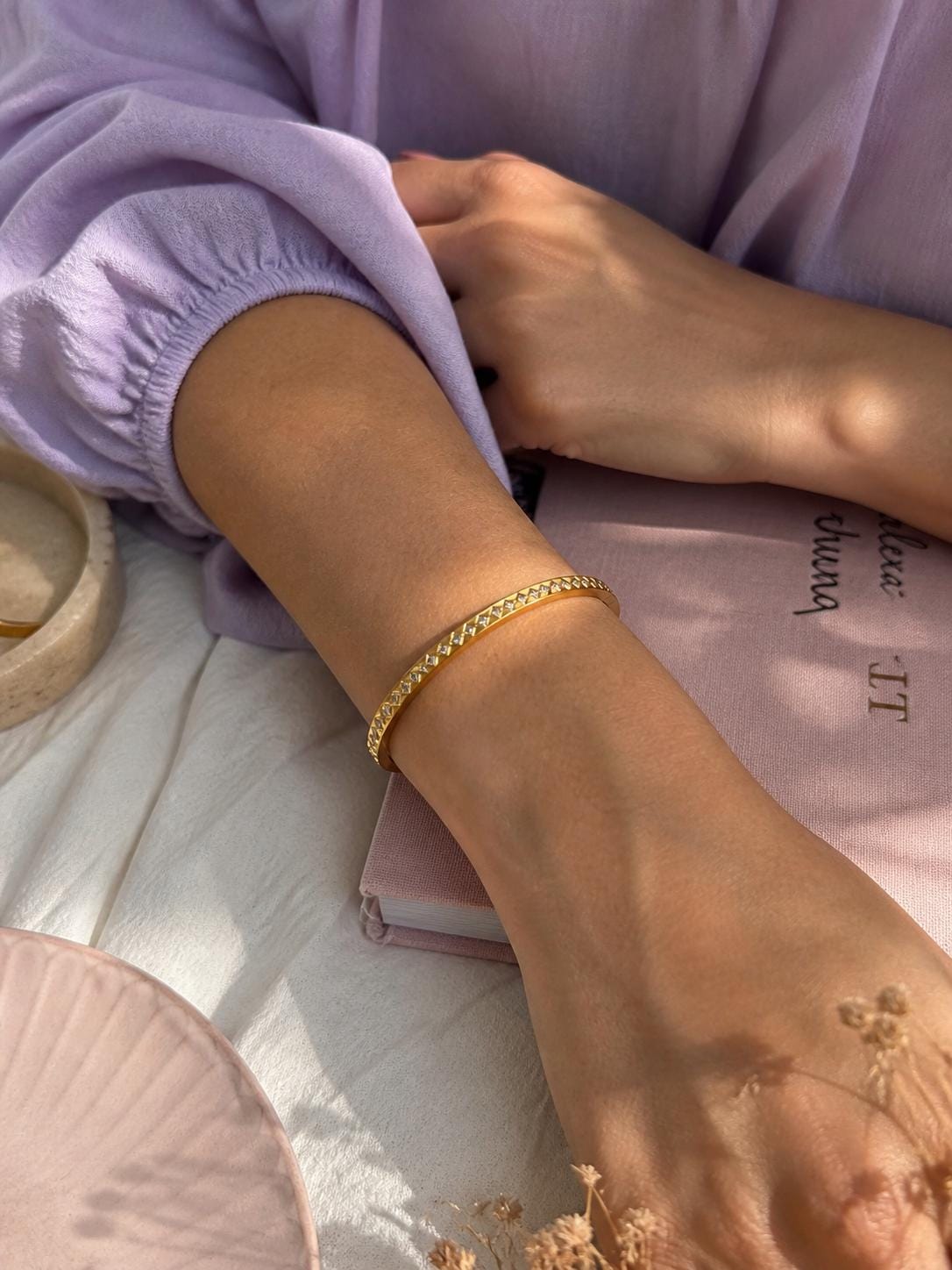 A gold-colored bangle bracelet with a repeating pattern of small, sparkling embellishments. The bracelet is worn on a person's arm, with a soft purple sleeve visible in the background. The bracelet is a jewelry item.