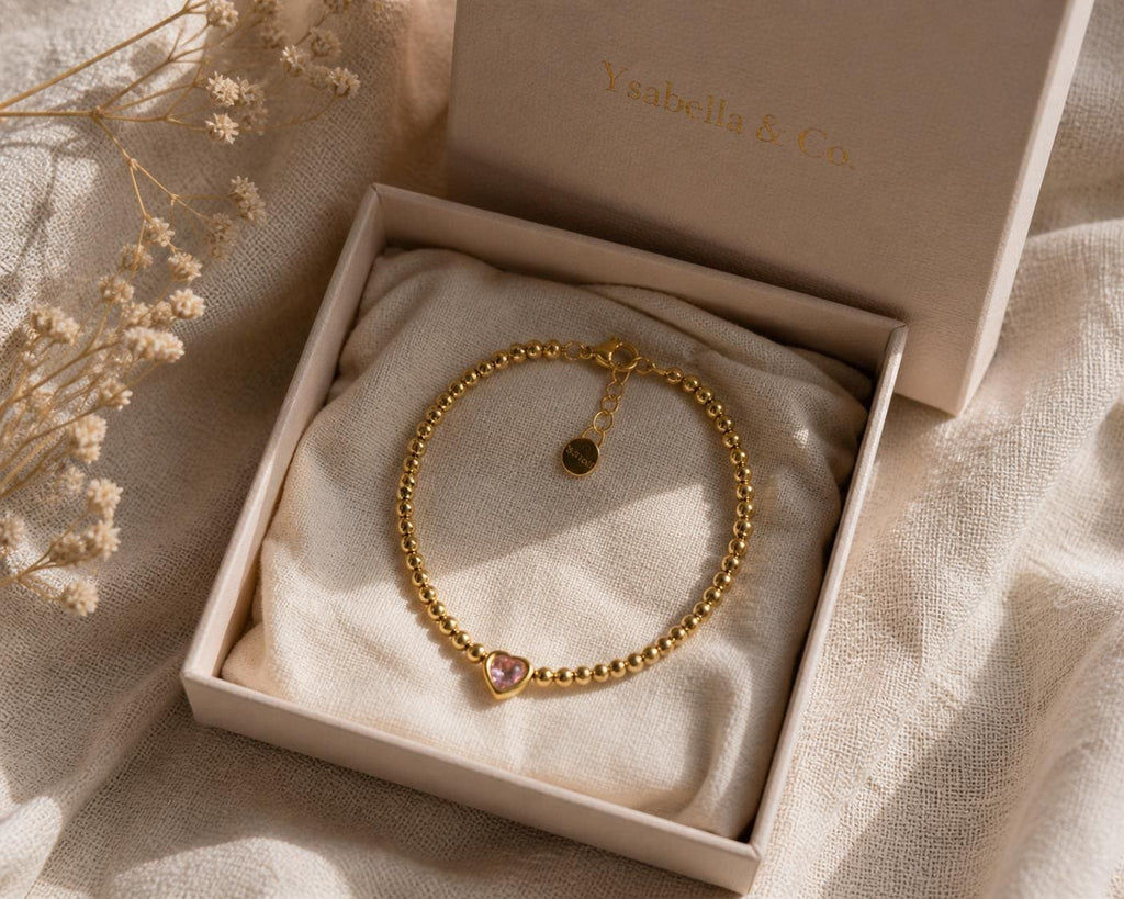 A gold beaded bracelet with a pink heart-shaped charm, presented in a light pink box. The box is open, revealing the bracelet resting on a cream-colored fabric. The brand name "Ysaabella & Co." is printed on the box.