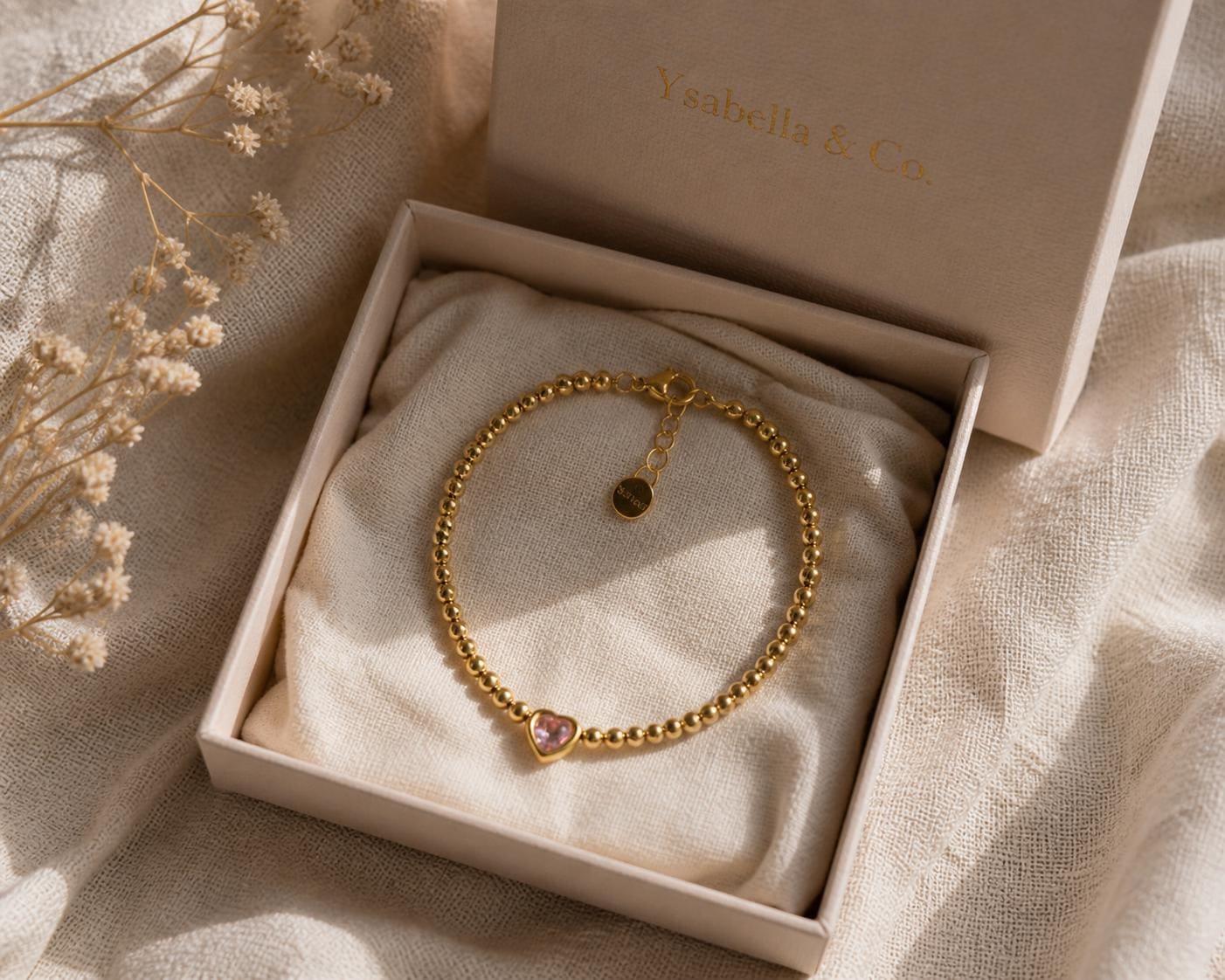 A gold beaded bracelet with a pink heart-shaped charm, presented in a light pink box. The box is open, revealing the bracelet resting on a cream-colored fabric. The brand name "Ysaabella & Co." is printed on the box.
