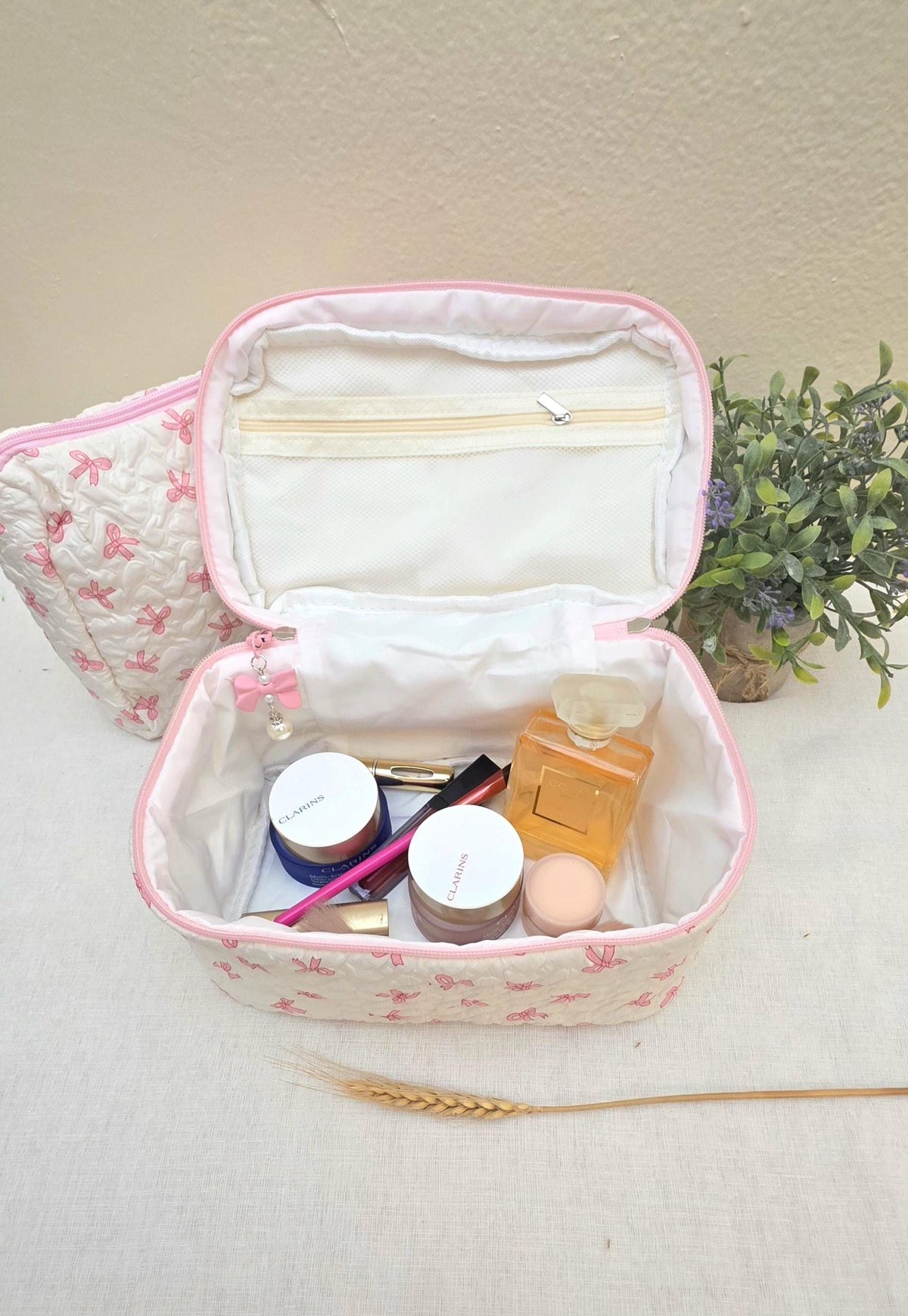 Image shows an Open pink and white quilted cosmetic bag with a bow pattern, filled with makeup products. The bag has a zipper closure and a small bow charm. A smaller matching bag is visible in the background.