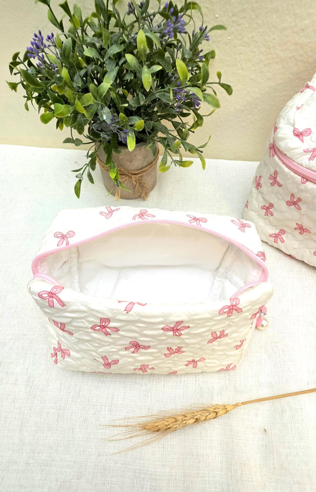 White quilted cosmetic bag with a pink bow pattern and a pink zipper. The bag is open, revealing a white interior. A matching backpack is visible in the background. A small potted plant is in the background.