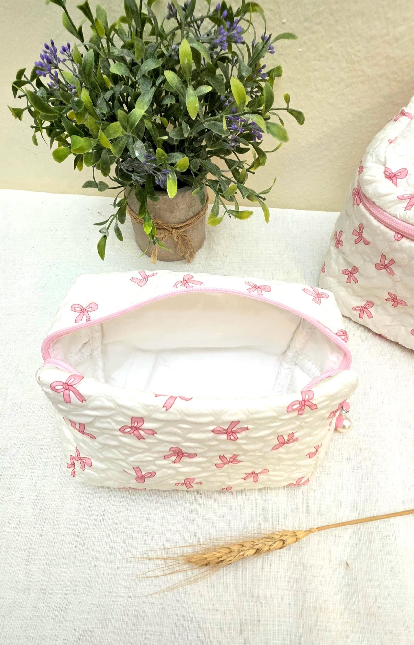 White quilted cosmetic bag with a pink bow pattern and a pink zipper. The bag is open, revealing a white interior. A matching backpack is visible in the background. A small potted plant is in the background.
