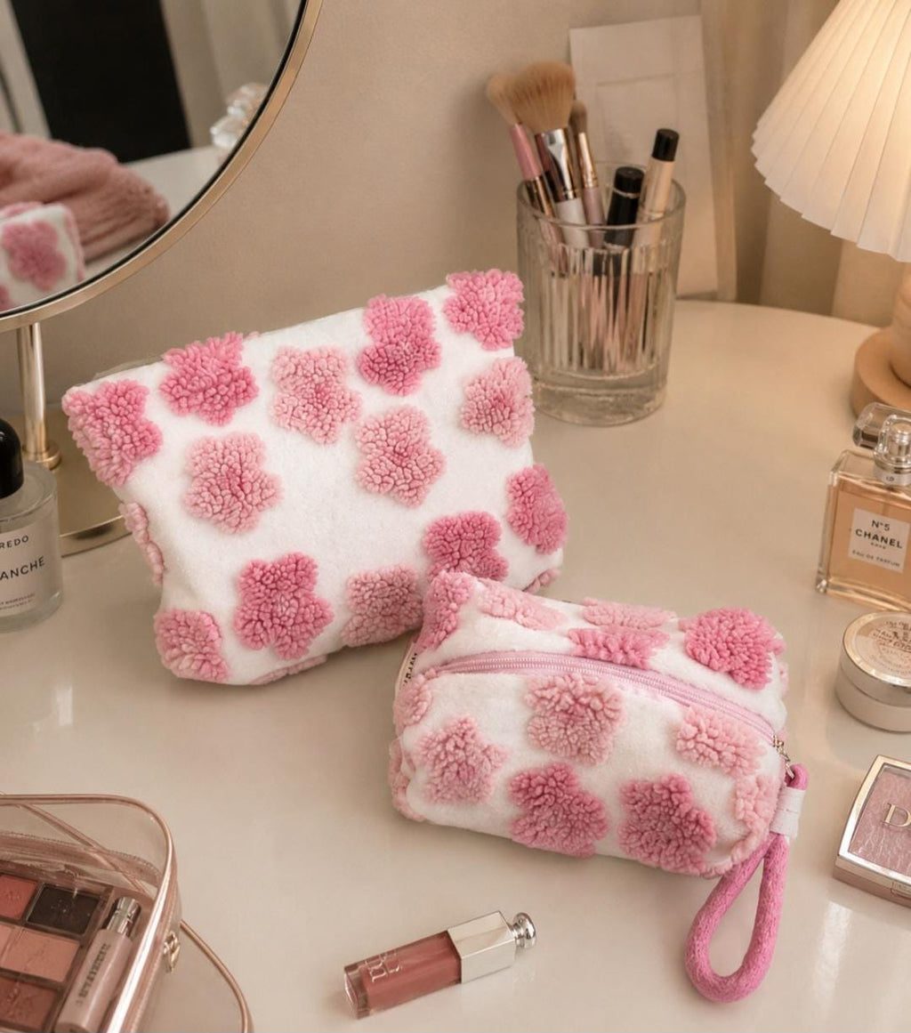 Two white cosmetic bags with pink flower-shaped accents. One bag is rectangular, and the other is smaller with a zipper and a pink wrist strap. Makeup brushes and perfume bottles are in the background. Cosmetic / makeup bag in 2 designs : handbag and clutch .
