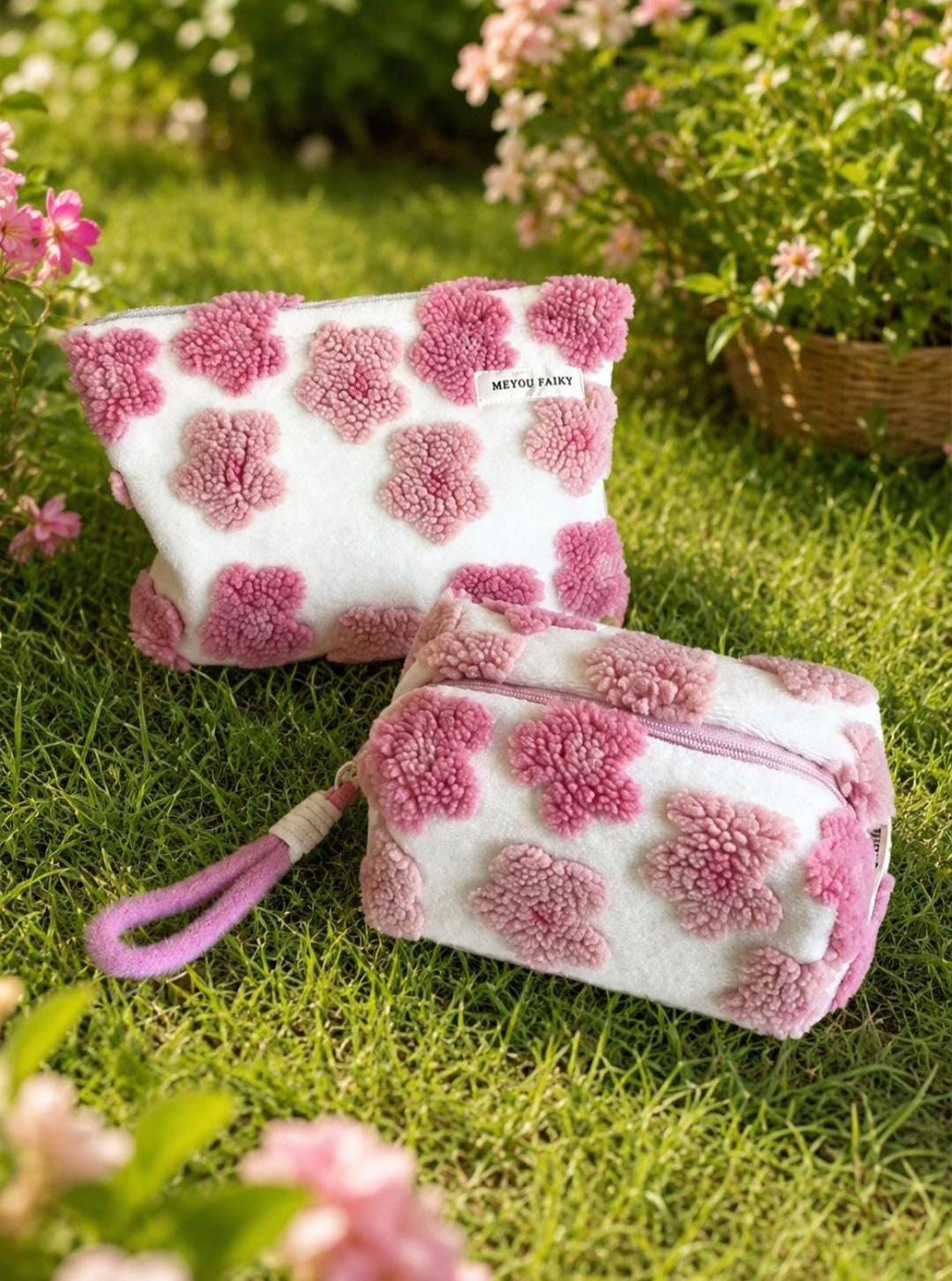 Two white cosmetic bags with a pink floral pattern. One bag is open, and the other has a zipper and a pink handle. The bags are on a bed of green grass, with pink flowers in the background. The text "MEYOU FAIKY" is on one of the bags.