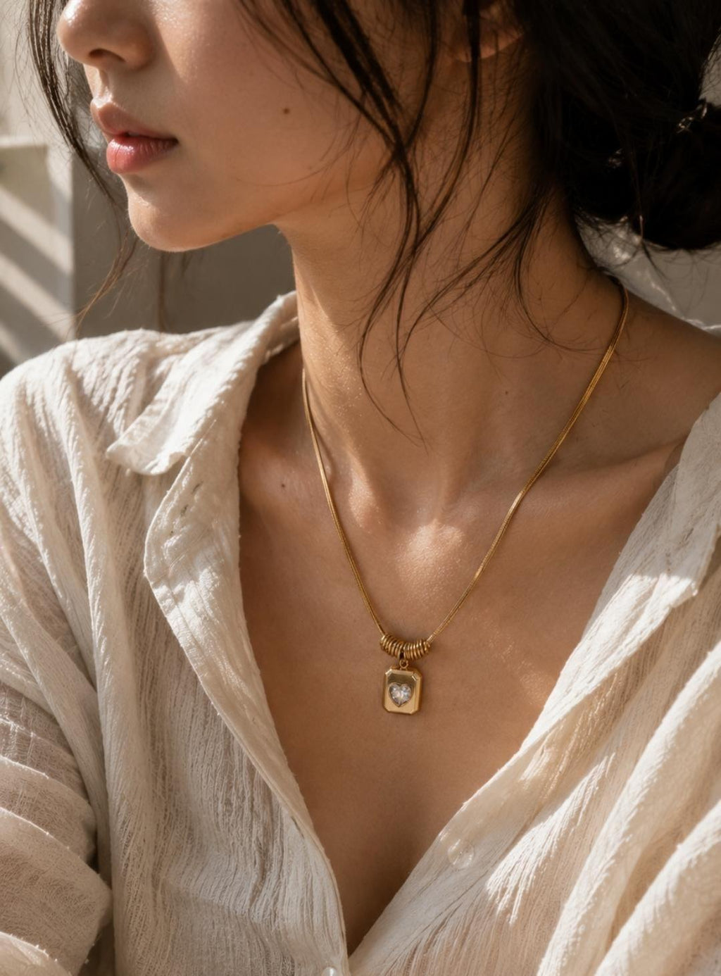 A gold-toned necklace featuring a flat snake chain and a rectangular pendant. The pendant has a central heart-shaped cutout with a clear stone. A small, ribbed cylindrical bead sits directly above the pendant on the chain.