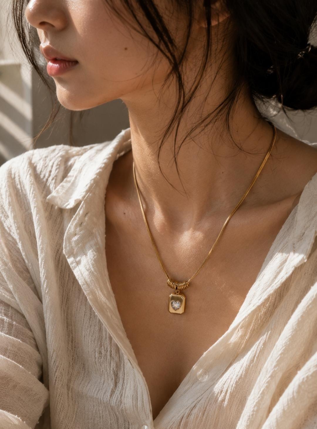 A gold-toned necklace featuring a flat snake chain and a rectangular pendant. The pendant has a central heart-shaped cutout with a clear stone. A small, ribbed cylindrical bead sits directly above the pendant on the chain.