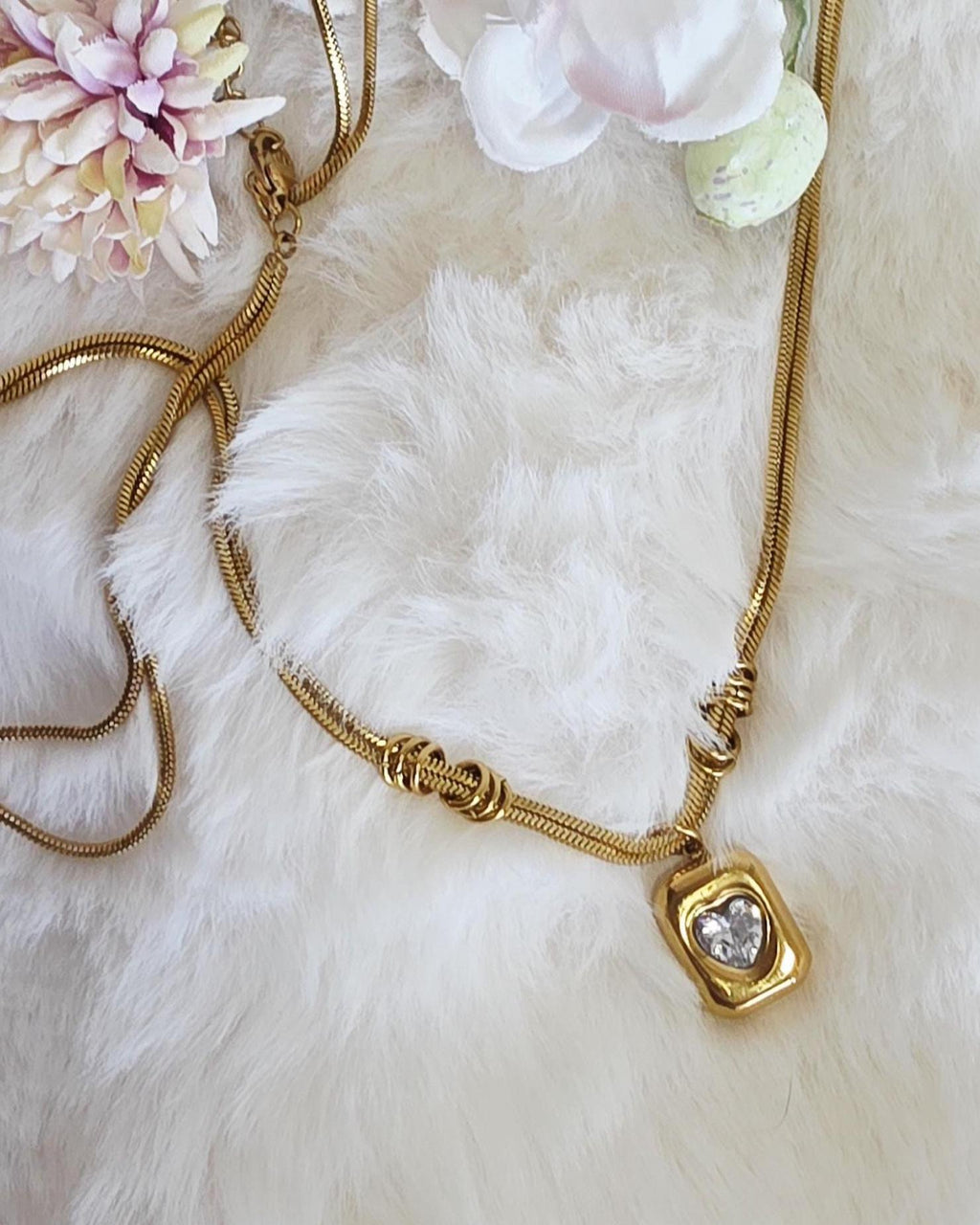 A gold-toned snake chain necklace featuring a rectangular pendant with a central heart-shaped clear stone. The chain includes two decorative gold-toned rings positioned along the length of the necklace near the pendant attachment point.