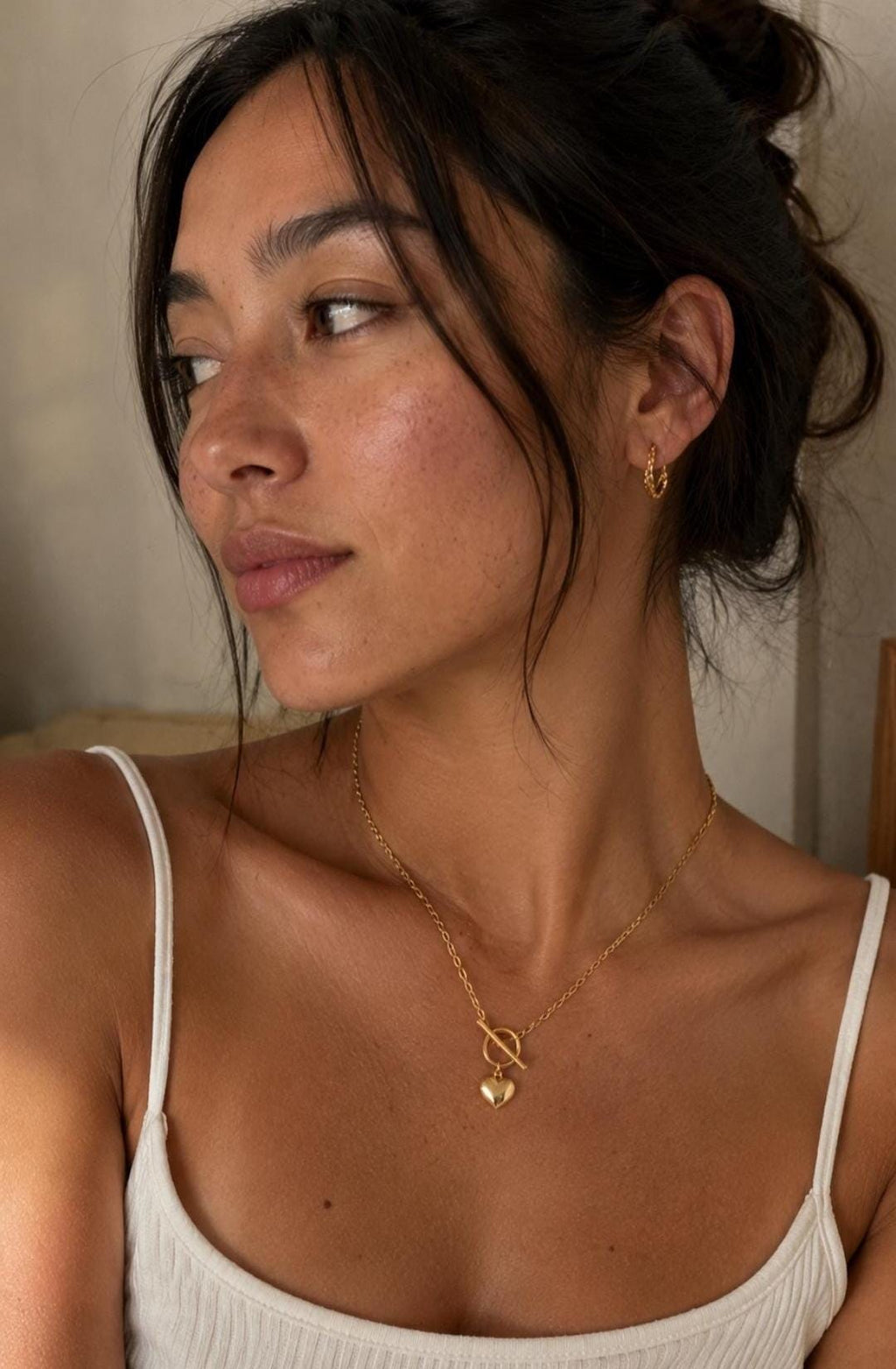 A gold-toned necklace featuring a delicate chain with a toggle clasp and a small, smooth heart-shaped pendant. The necklace is paired with a small, textured gold-toned hoop earring worn in the earlobe, completing the jewelry set.