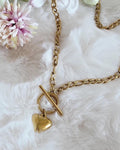 A gold-toned metal chain necklace featuring a toggle clasp closure and a smooth, polished heart-shaped pendant. The necklace consists of interlocking oval links that connect to a circular ring and a horizontal bar toggle mechanism.