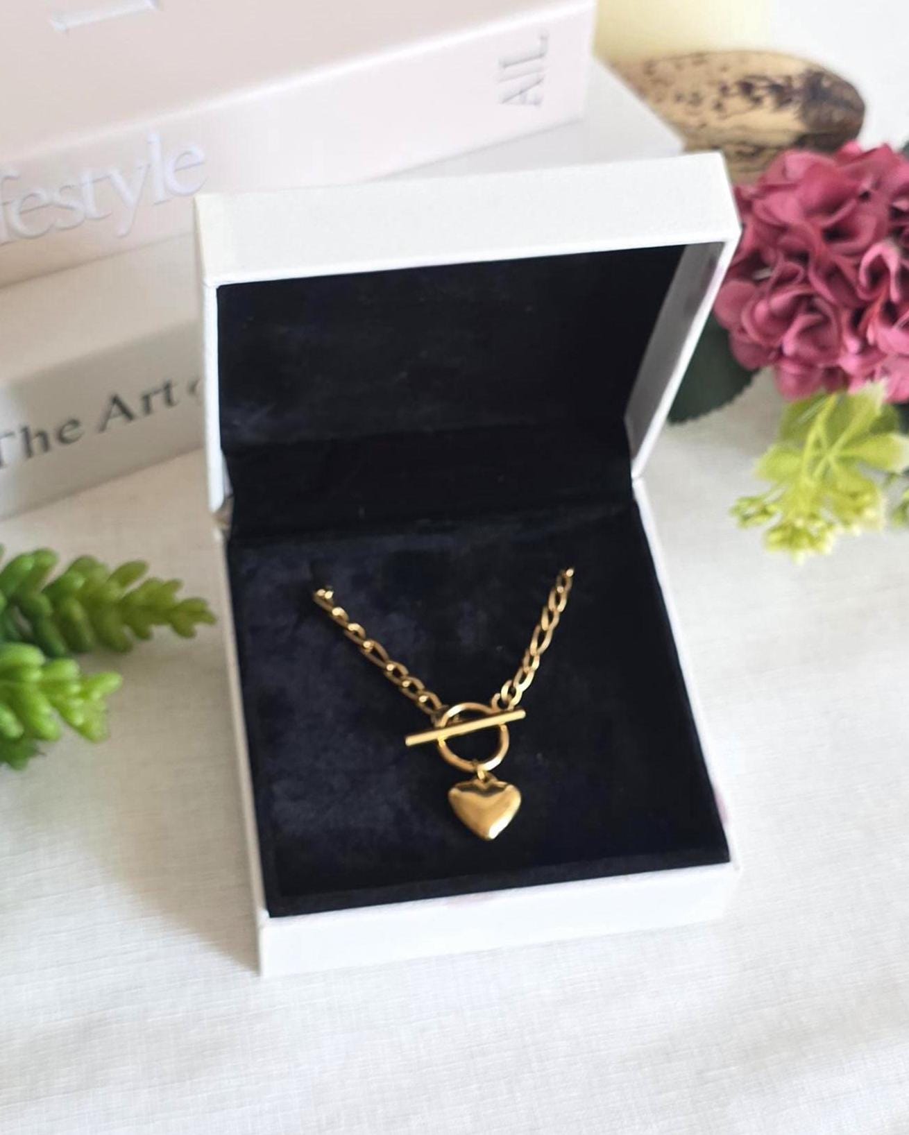 A gold-toned necklace featuring a thick chain, a toggle clasp, and a smooth heart-shaped pendant. The necklace is displayed inside an open white jewelry box with a black velvet interior lining, showcasing the metallic finish clearly.