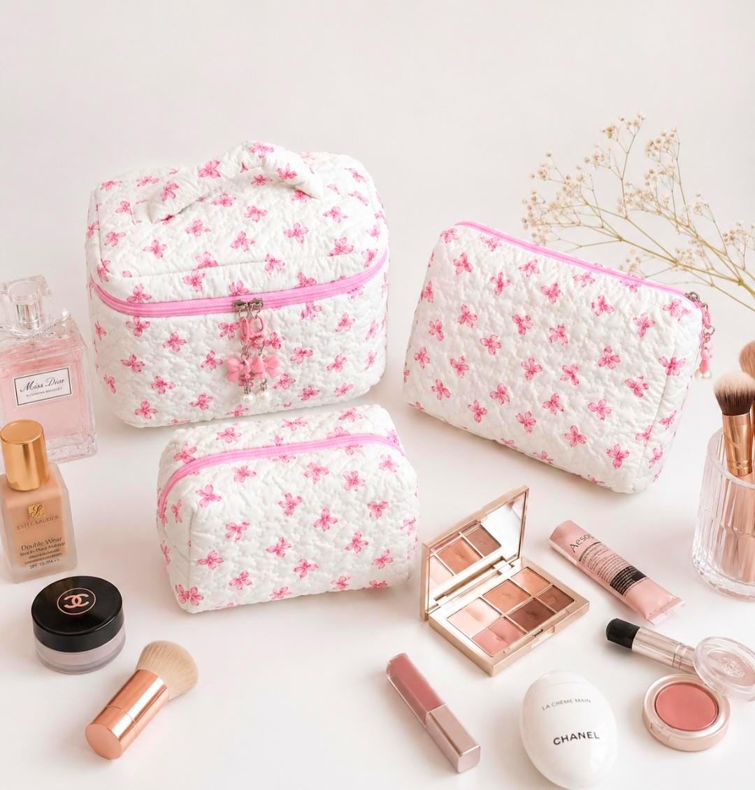 Image  includes : Three white quilted cosmetic bags with pink floral patterns and pink zippers. Makeup products, including a blush, lip gloss, and eyeshadow palette, are arranged around the bags. A perfume bottle and makeup brushes are also visible.