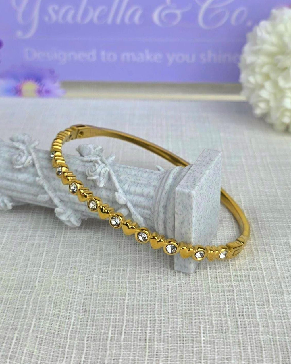 A gold-tone hinged bangle bracelet featuring a repeating pattern of small metal hearts alternating with clear, round-cut rhinestones. The bracelet has a polished metallic finish and a secure clasp mechanism visible on the side.