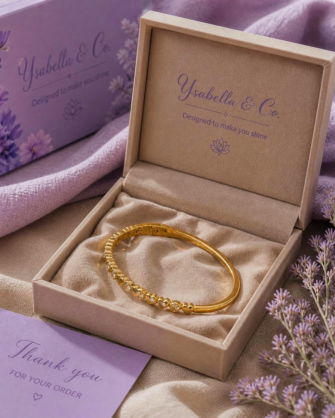 A gold-toned hinged bangle bracelet featuring a row of small heart-shaped links set with clear stones. The bracelet rests inside a beige velvet-lined jewelry box with the text Ysabella & Co. Designed to make you shine printed inside.
