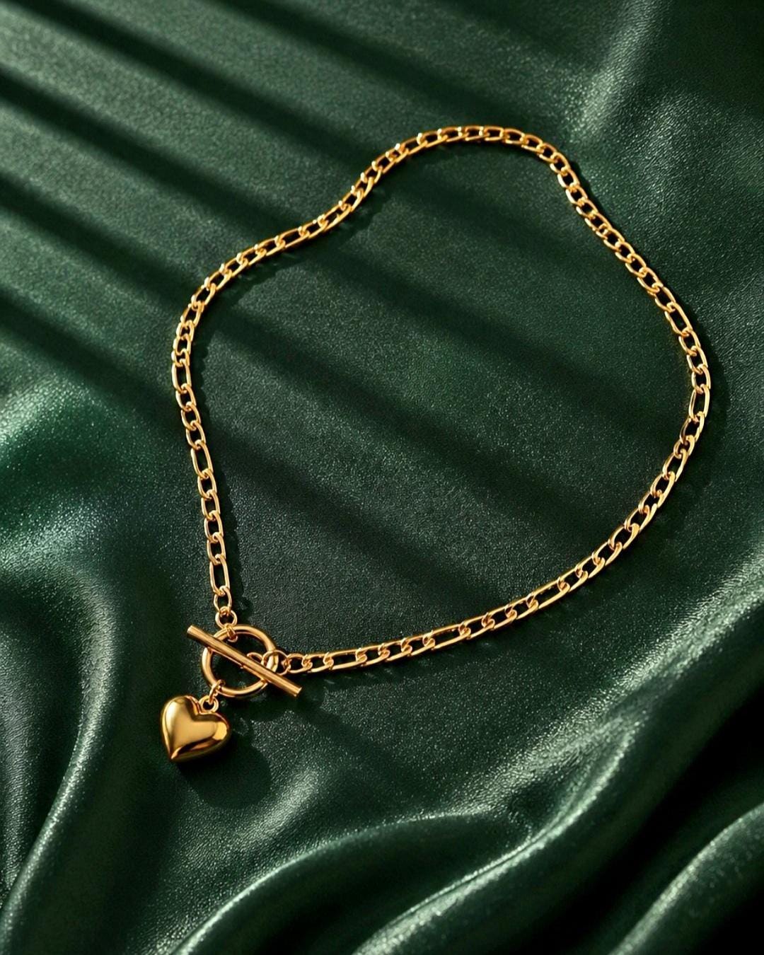 A gold-toned chain necklace featuring a toggle clasp closure and a smooth, polished heart-shaped pendant. The necklace is constructed with interlocking oval links and a central circular ring that secures the T-bar toggle mechanism.