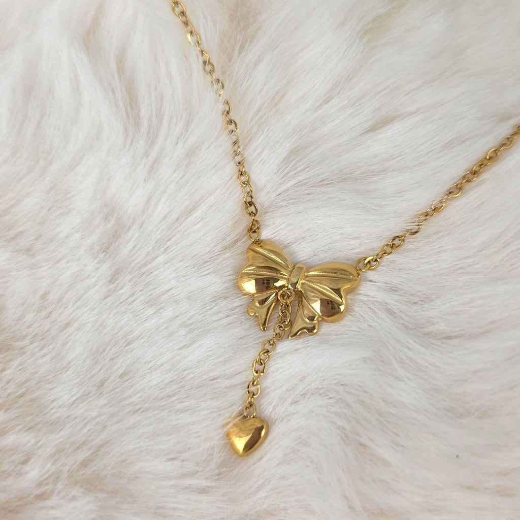 A gold-toned necklace featuring a central bow-shaped pendant with a polished finish. A delicate chain extends downward from the center of the bow, terminating in a small, smooth, heart-shaped charm that hangs as a drop element.
