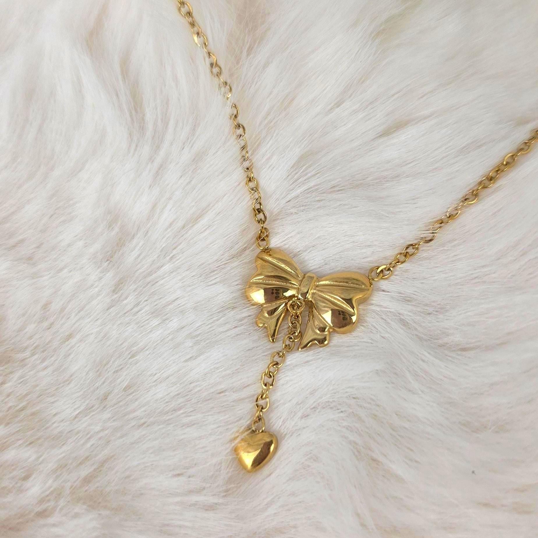 A gold-toned necklace featuring a central bow-shaped pendant with a polished finish. A delicate chain extends downward from the center of the bow, terminating in a small, smooth, heart-shaped charm that hangs as a drop element.
