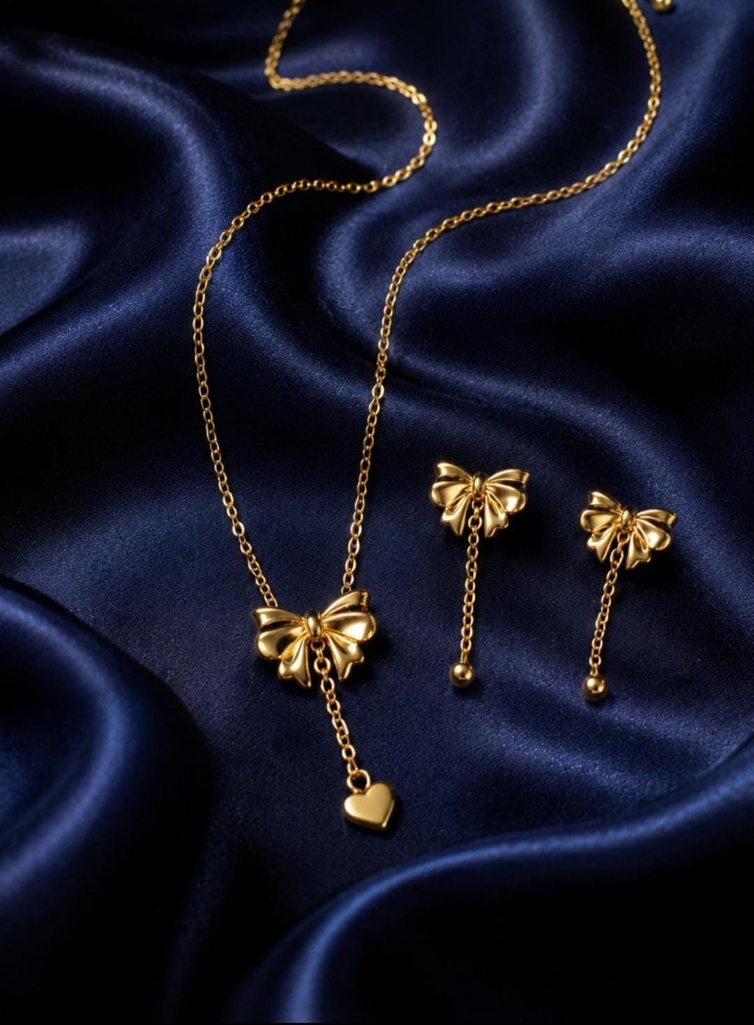 This image has A gold-toned jewelry set consisting of a necklace and two stud earrings. The necklace features a bow pendant with a dangling heart charm. Each earring consists of a bow stud with a short chain ending in a small gold sphere. 18k Gold plated jewelry.