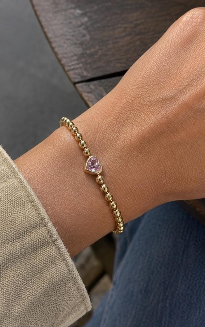 Image includes a  Gold beaded bracelet with a pink heart-shaped charm. The bracelet is worn on a wrist, with a cream-colored sleeve in the background. The bracelet is made of small, round gold beads and the heart charm is a light pink color.