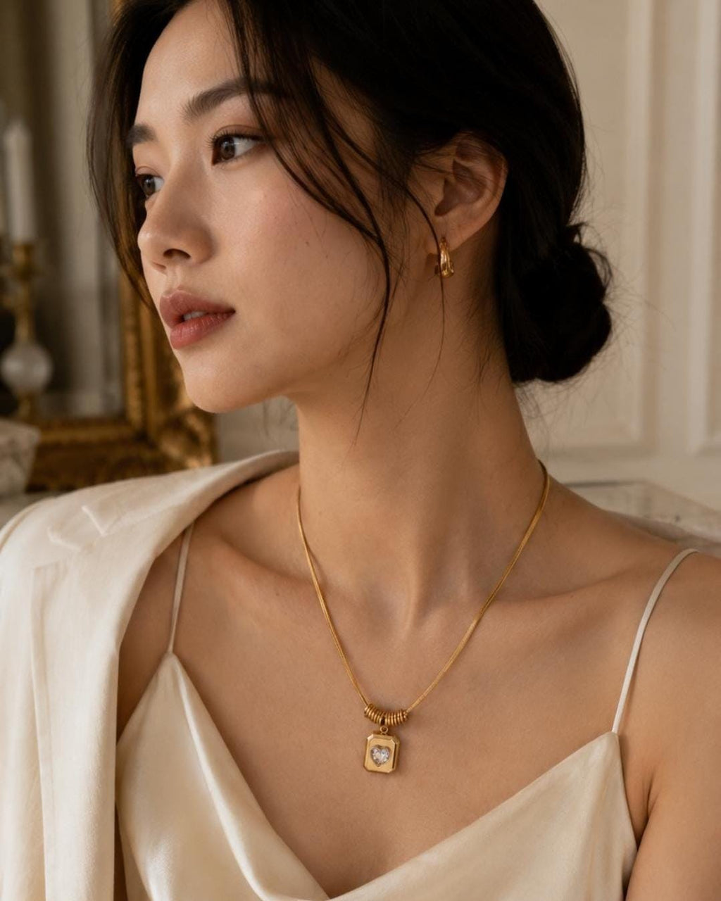 A gold-toned necklace featuring a thin snake chain and a rectangular pendant with a central heart-shaped cutout containing a clear stone. The pendant is suspended from a small ribbed cylindrical bead. A matching gold-toned hoop earring is visible.