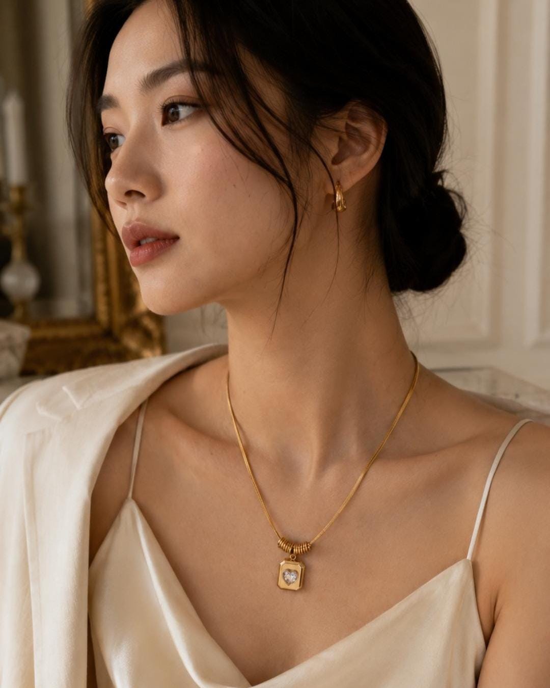 A gold-toned necklace featuring a thin snake chain and a rectangular pendant with a central heart-shaped cutout containing a clear stone. The pendant is suspended from a small ribbed cylindrical bead. A matching gold-toned hoop earring is visible.