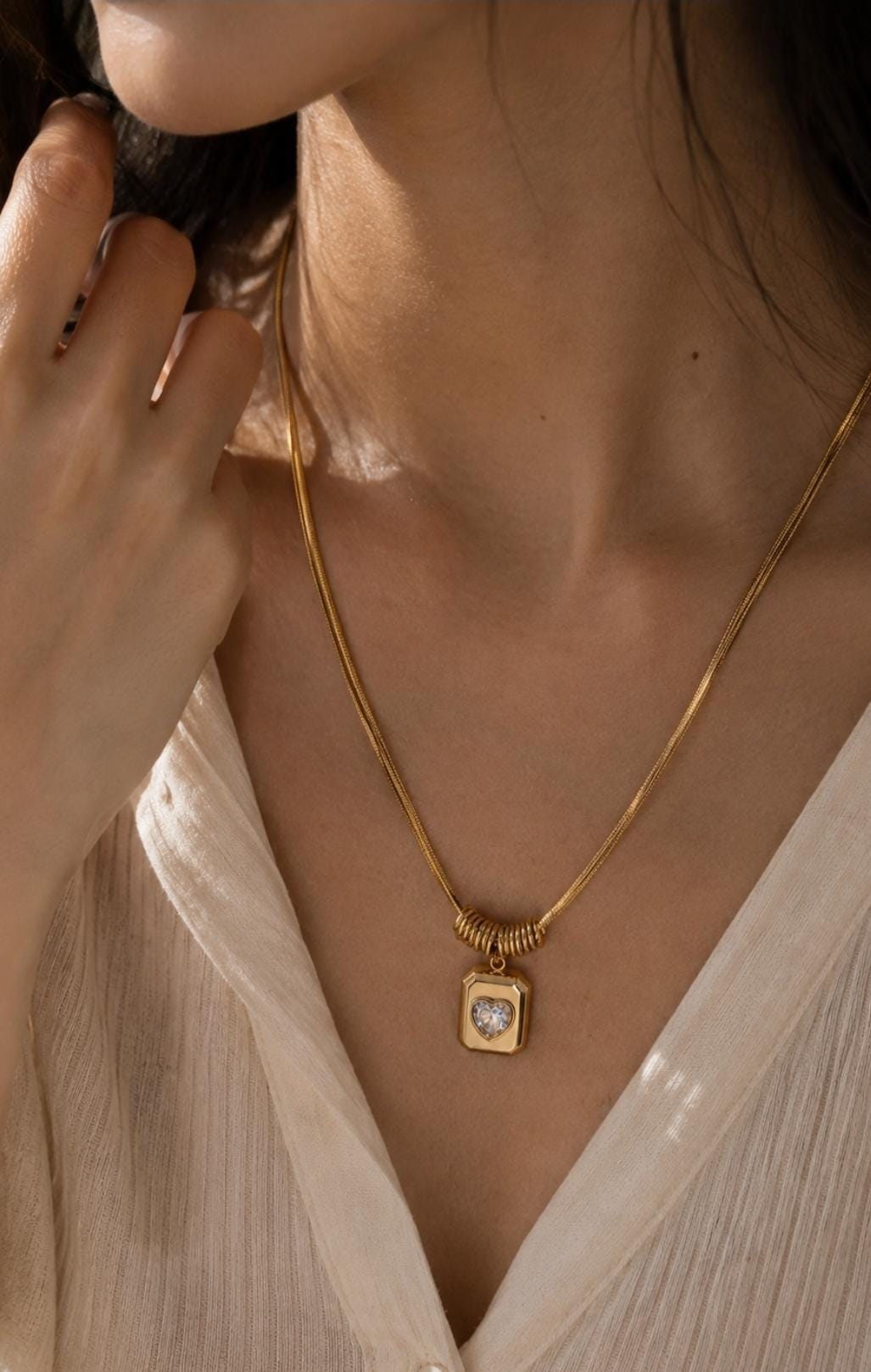 A gold-toned snake chain necklace featuring a rectangular pendant with a central heart-shaped clear stone. The pendant is suspended from a series of small gold-toned rings that slide along the chain, creating a layered, textured appearance.