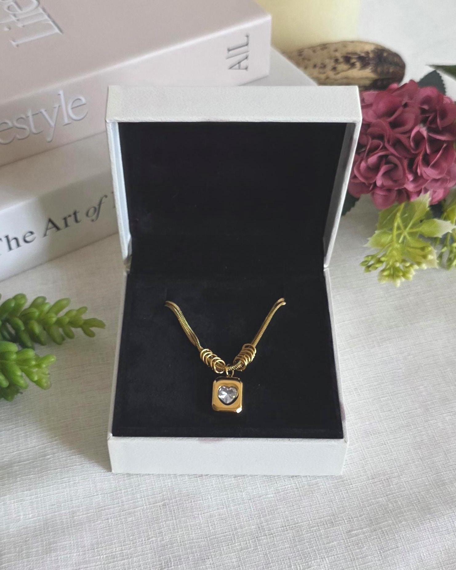 A gold-toned necklace featuring a rectangular pendant with a central heart-shaped clear stone, suspended from a chain with multiple circular links near the charm, presented inside a white box with a black velvet interior lining.