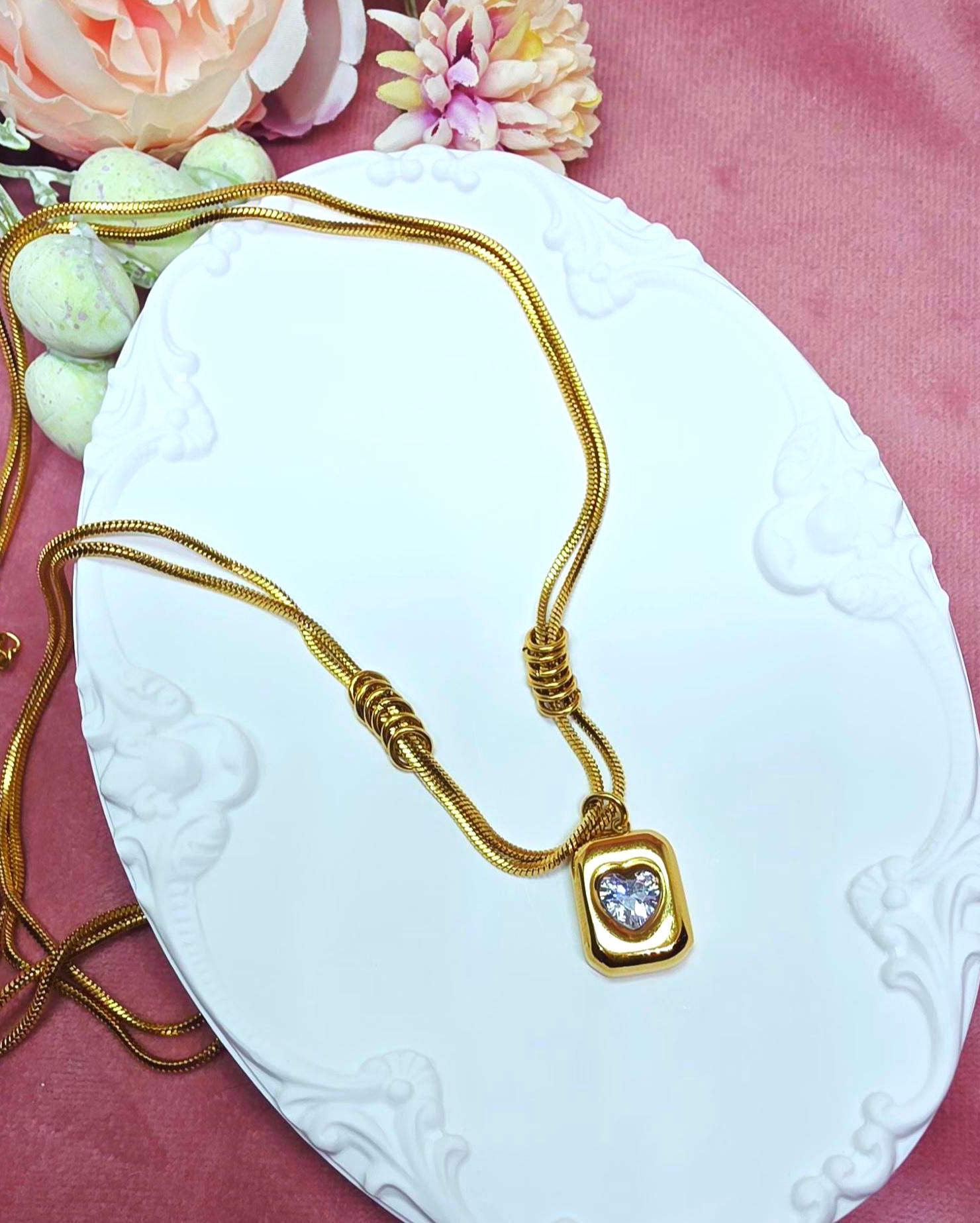 A gold-toned snake chain necklace featuring a rectangular pendant with a central heart-shaped clear stone. The chain includes two sets of three decorative gold rings positioned above the pendant, creating a layered, structured look.