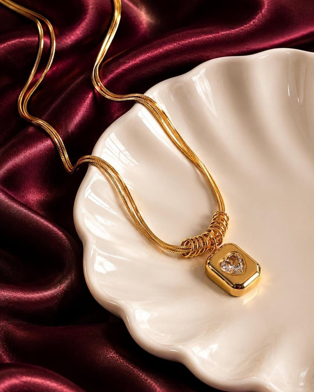 A gold-toned necklace featuring a double snake chain that passes through a series of small rings, leading to a rectangular pendant with rounded edges and a central heart-shaped clear gemstone set within the smooth metal surface.