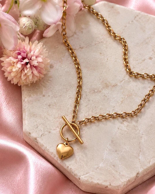 A gold-toned chain necklace featuring a toggle clasp closure and a small, smooth heart-shaped pendant. The necklace is constructed with oval-shaped links and has a polished metallic finish, showcasing a classic design with a central heart charm.