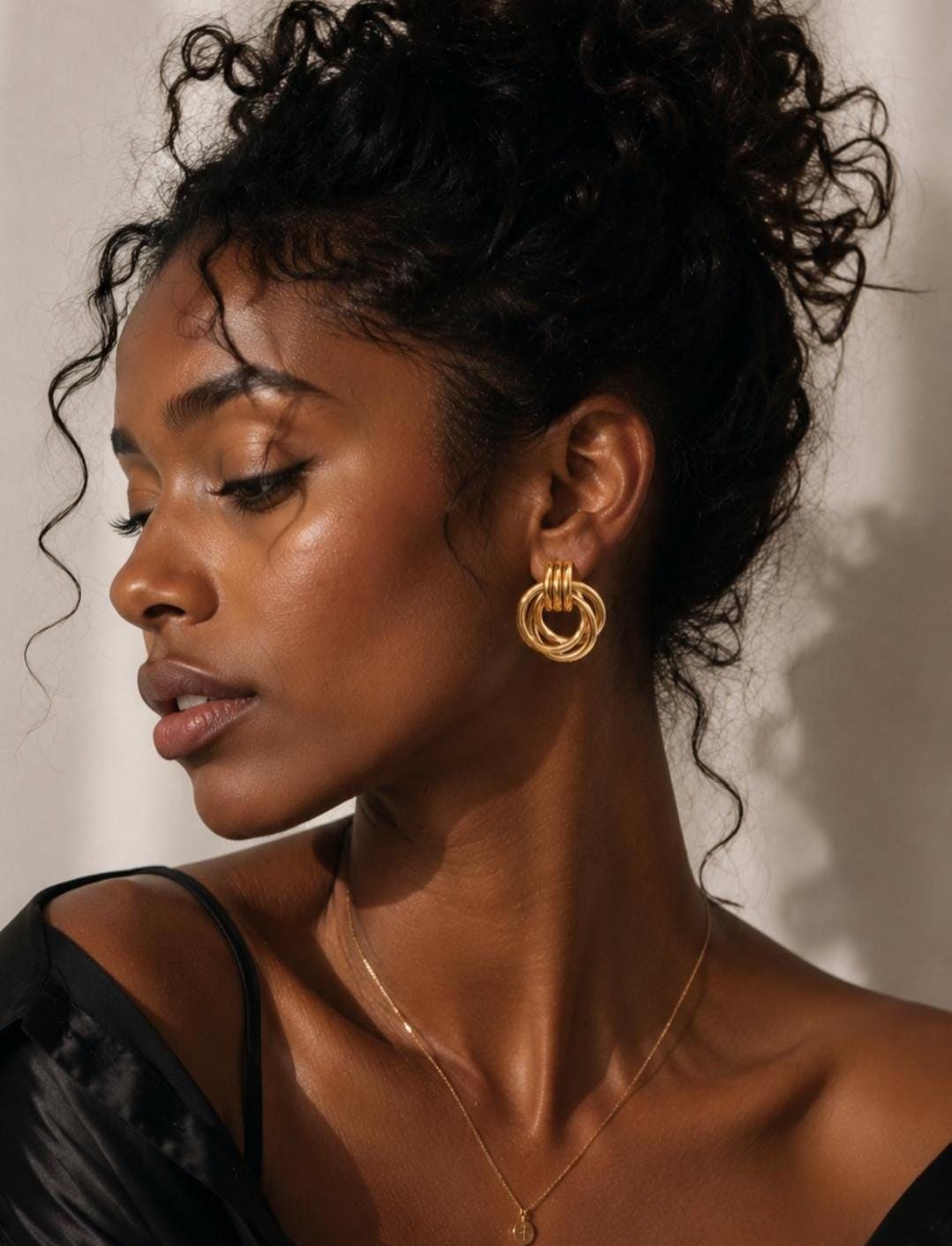A pair of gold-toned earrings featuring three interlocking circular rings that hang from a small, solid top loop. The earrings have a polished, metallic finish and are worn on the earlobe, complemented by a thin gold-toned chain necklace.