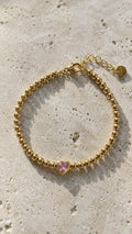 Image shows aGold beaded bracelet with a pink heart-shaped charm on a  beige stone background.