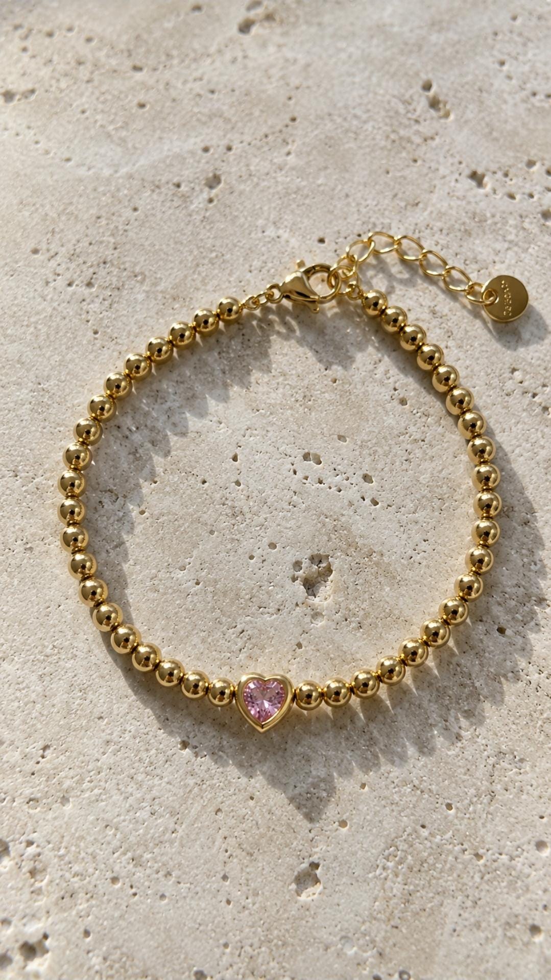 Image shows aGold beaded bracelet with a pink heart-shaped charm on a  beige stone background.