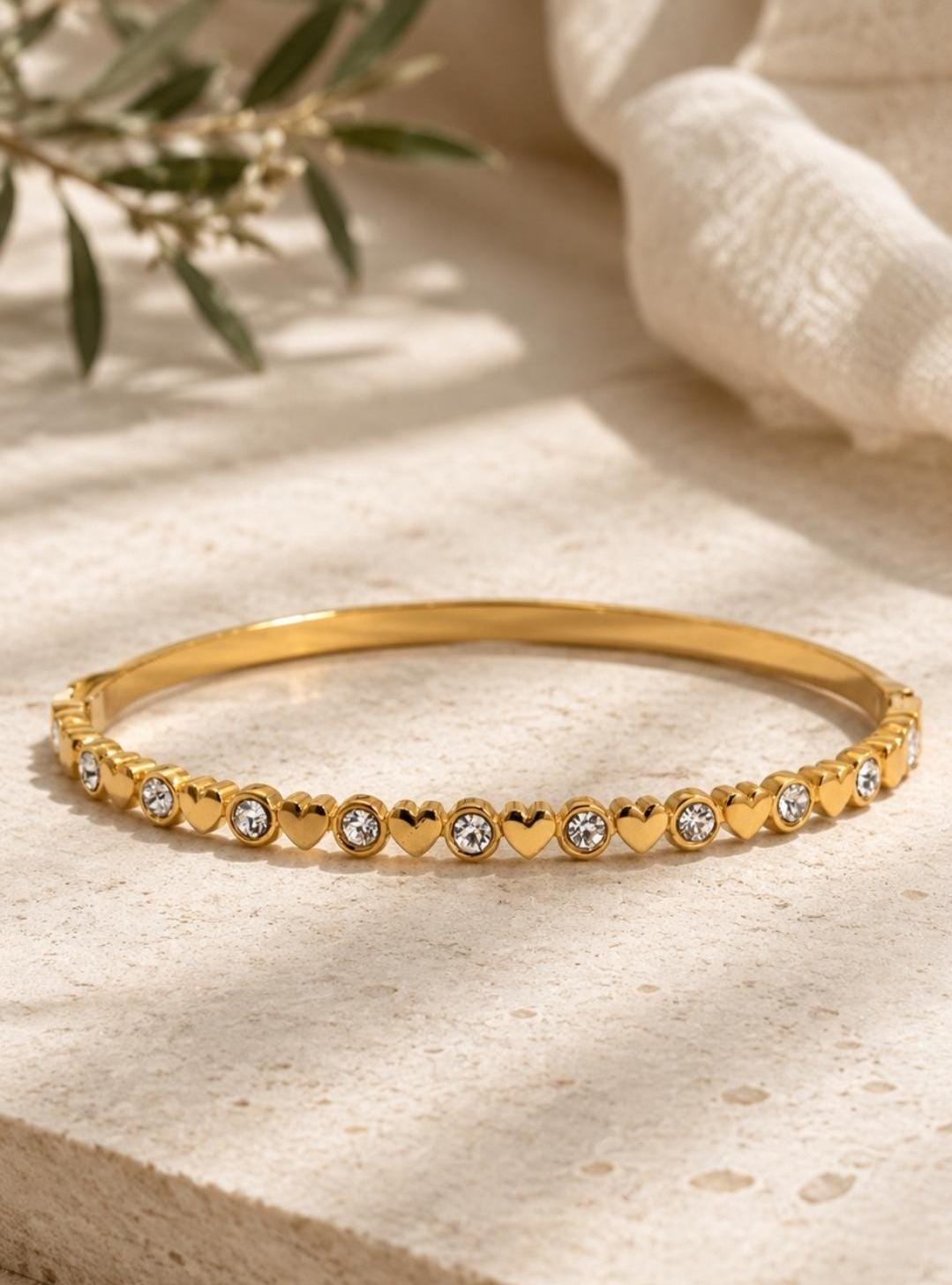 A gold-toned metal bangle bracelet featuring an alternating pattern of small, polished heart shapes and round, clear, sparkling crystals set along the entire outer circumference of the thin, circular band.