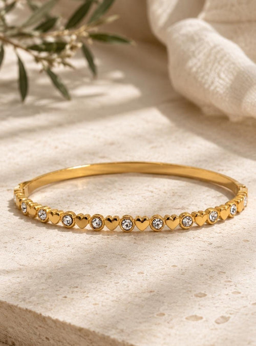 A gold-toned metal bangle bracelet featuring an alternating pattern of small, polished heart shapes and round, clear, sparkling crystals set along the entire outer circumference of the thin, circular band.