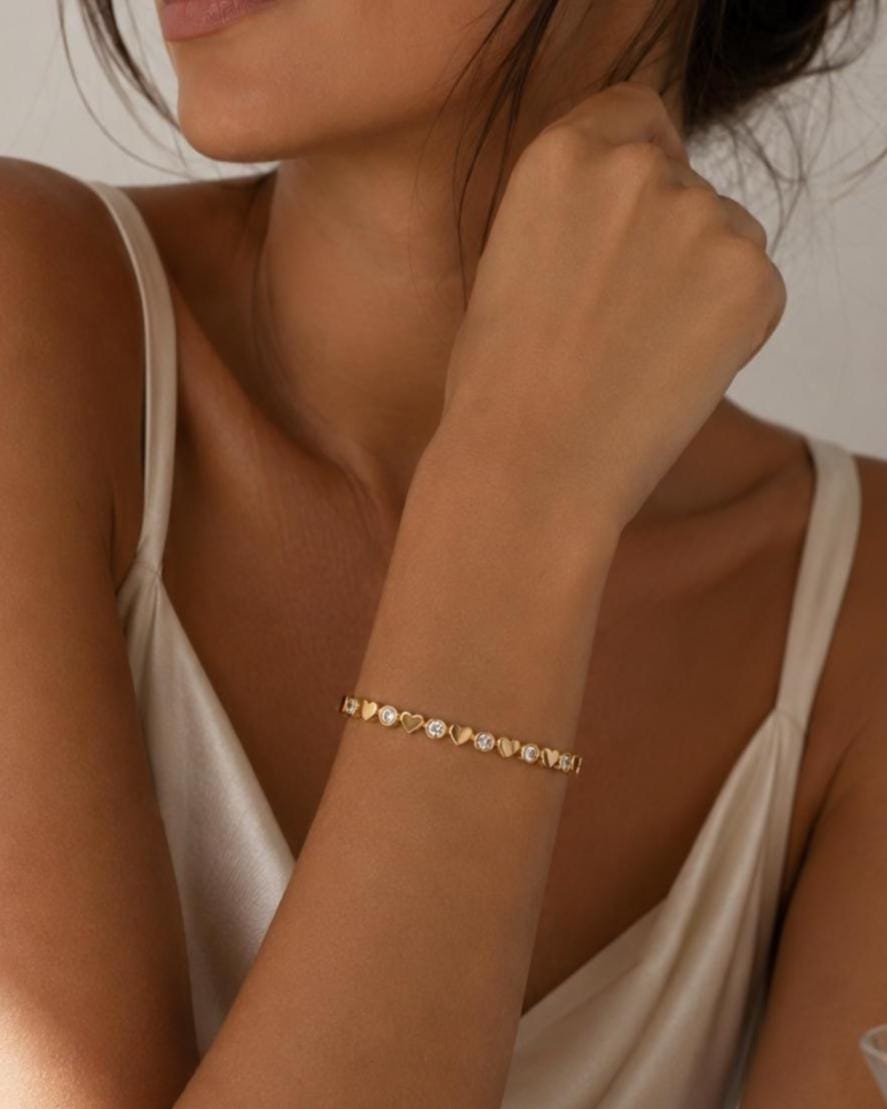 A gold-toned bracelet featuring an alternating pattern of small, solid gold hearts and round, clear, sparkling stones. The delicate chain links these decorative elements together in a continuous, symmetrical design around the wrist.