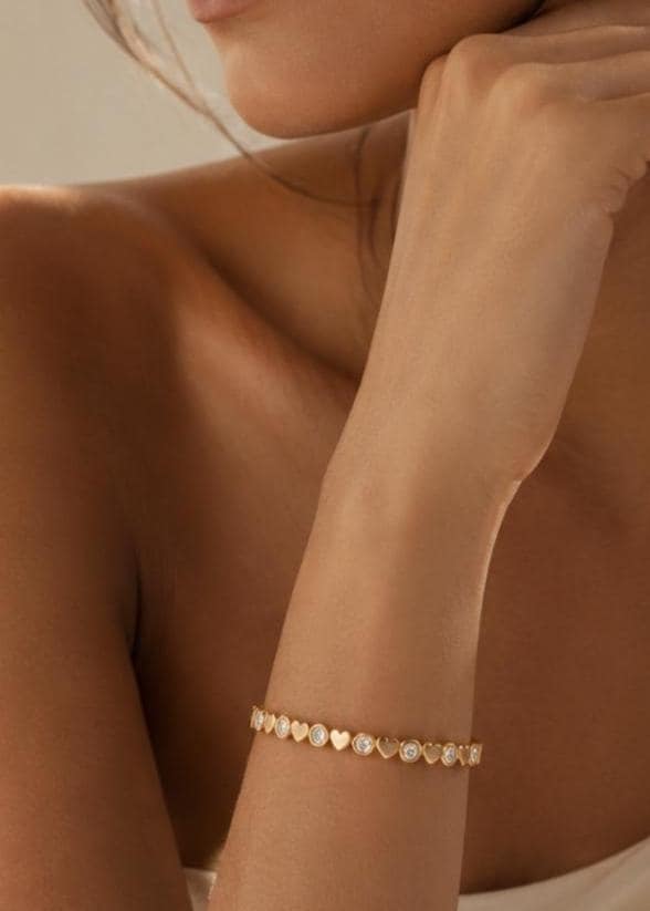 A gold-toned bracelet featuring an alternating pattern of small, solid heart-shaped links and circular bezel-set clear stones. The jewelry piece wraps around the wrist, showcasing a repeating design of metallic hearts and sparkling round accents.