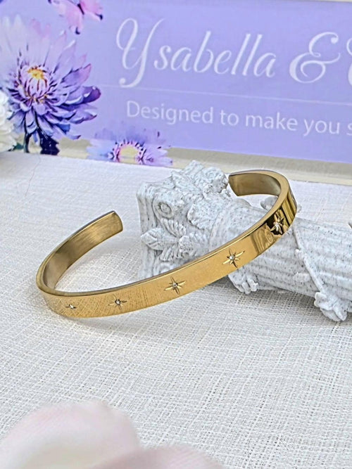 May include: Gold-toned cuff bracelet with star accents. The open-ended bracelet features a brushed metal finish and small, sparkling star-shaped embellishments. The background includes a sign that says "Ysabella & Co. Designed to make you smile."