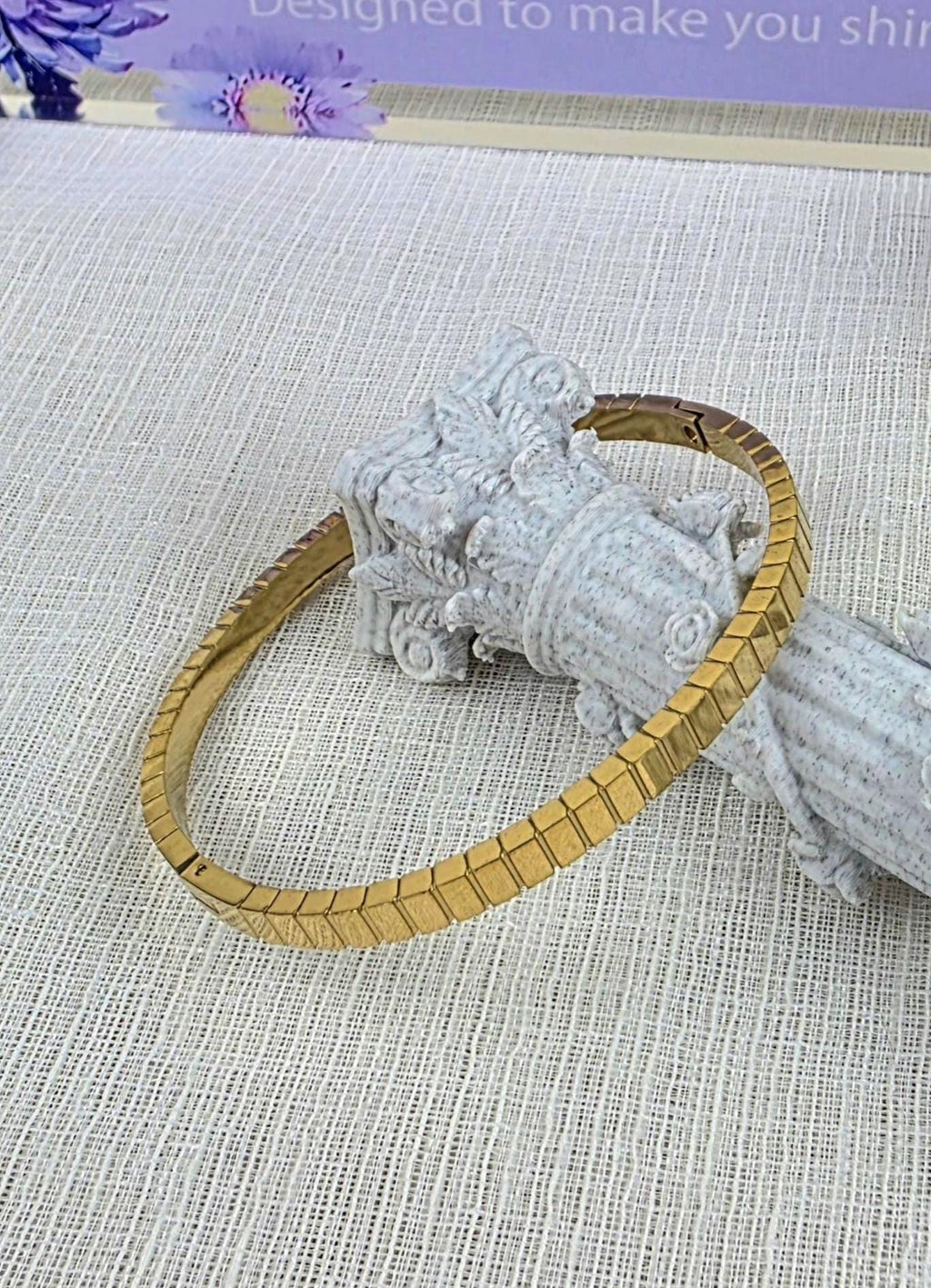 A gold-toned metal bracelet featuring a series of interconnected, rectangular, chevron-patterned links. The bracelet has a smooth, polished finish and a hinged clasp mechanism, displayed resting against a textured, light-colored column.