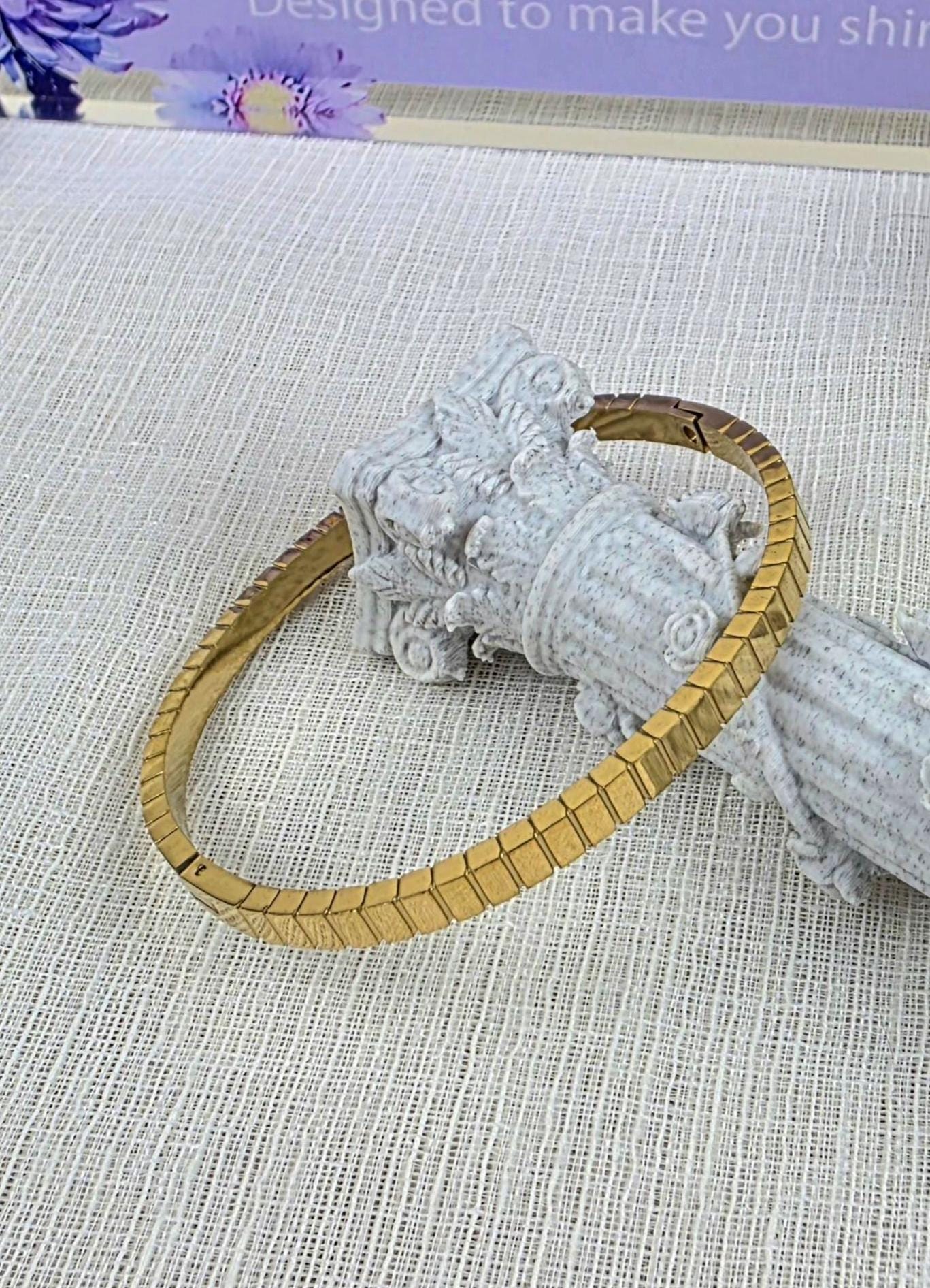 A gold-toned metal bracelet featuring a series of interconnected, rectangular, chevron-patterned links. The bracelet has a smooth, polished finish and a hinged clasp mechanism, displayed resting against a textured, light-colored column.