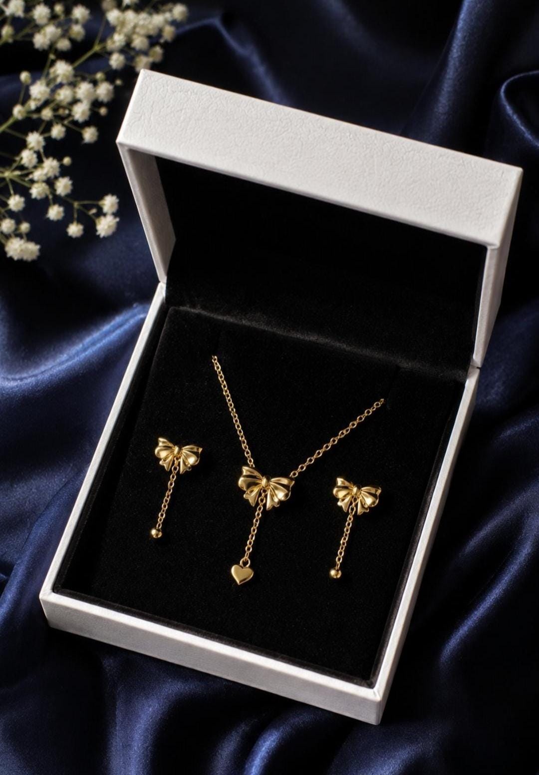 This photo  includes A gold-toned jewelry set consisting of a necklace and two earrings presented in a white box. The necklace features a bow pendant with a dangling heart charm. Each earring features a bow design with a delicate chain and a small bead.